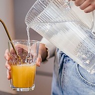 Ein Glas mit orangefarbenem Saft und Metallstrohhalm wird mit Wasser aus einer Karaffe aufgegossen.