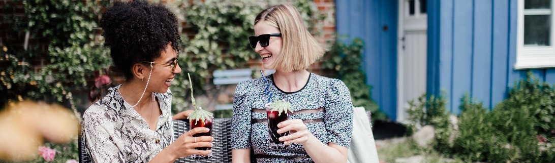 Zwei lachende Frauen mit einem Glas Sommerbowle sitzen auf einer Gartencouch